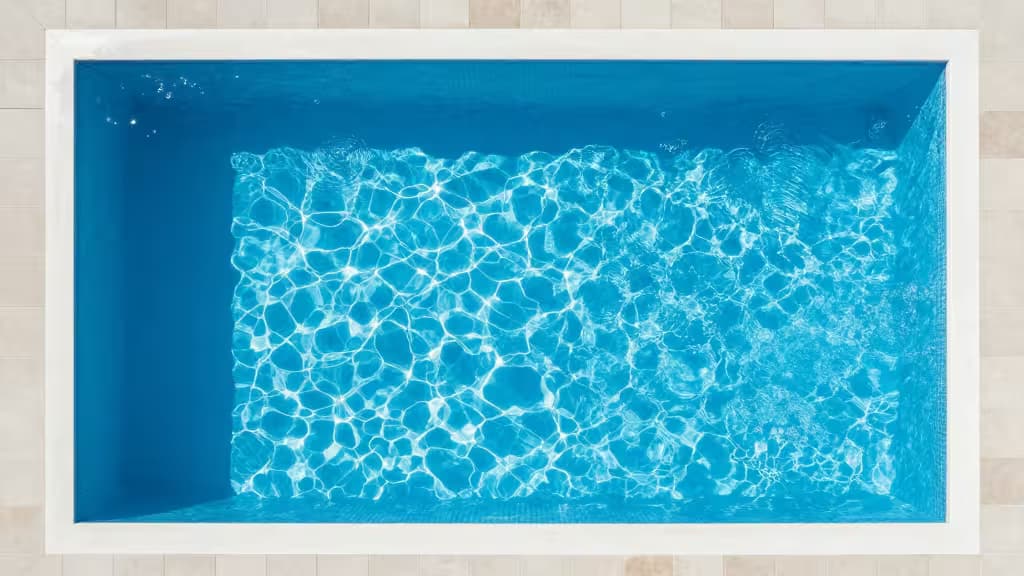 Rechthoekig zwembad met witte rand, blauw water en zonlicht rimpels op beige tegelomgeving.