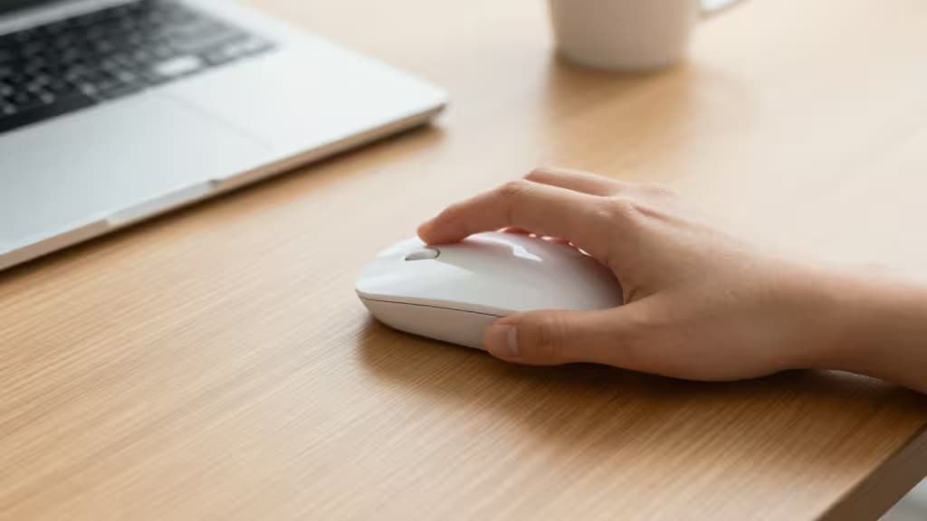 A small hand grips a white wireless mouse on a light wood desk beside a laptop.