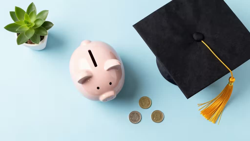Pink piggy bank, graduation cap, coins, and succulent on light blue background.