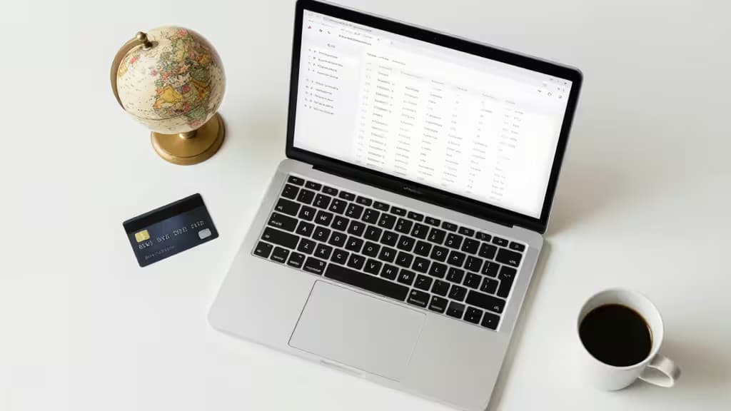 Laptop displays e-commerce dashboard beside globe, credit card, and coffee cup on white desk.