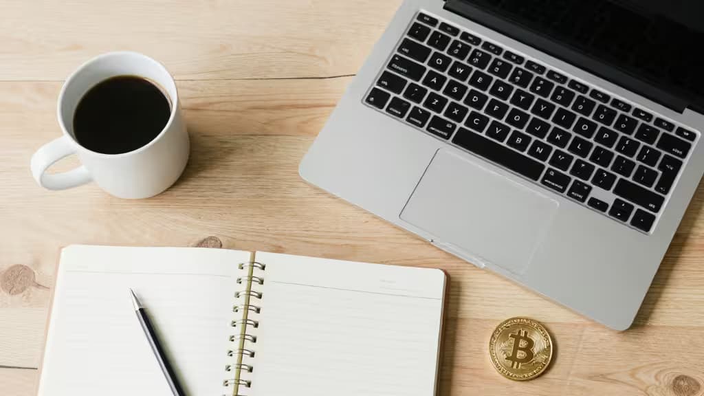Una laptop, libreta, taza de café y una moneda de Bitcoin sobre un escritorio de madera.