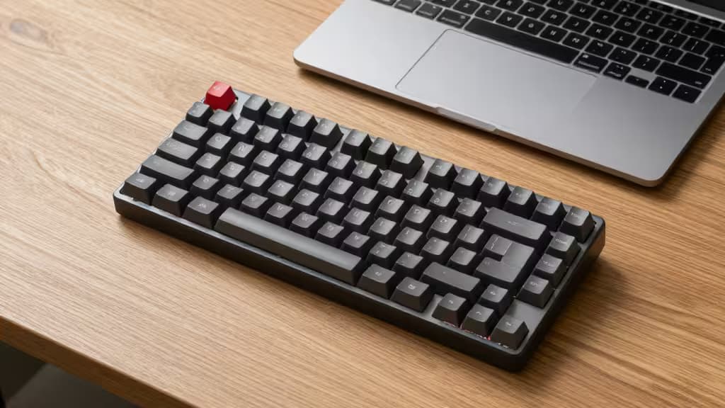 Low-profile mechanical keyboard with red escape key sits beside MacBook on wood desk.