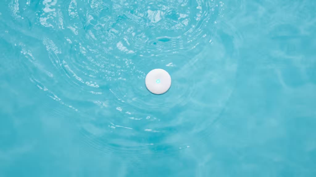 Top-down view of a calm swimming pool with a small floating smart water sensor device glowing with a soft blue indicator light in clear turquoise water.