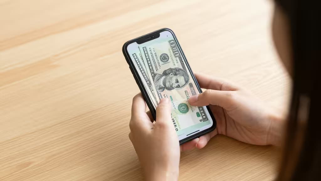 Persona sosteniendo un teléfono móvil junto a billetes de dólar y peso filipino sobre una mesa de madera, representando el envío de remesas internacionales