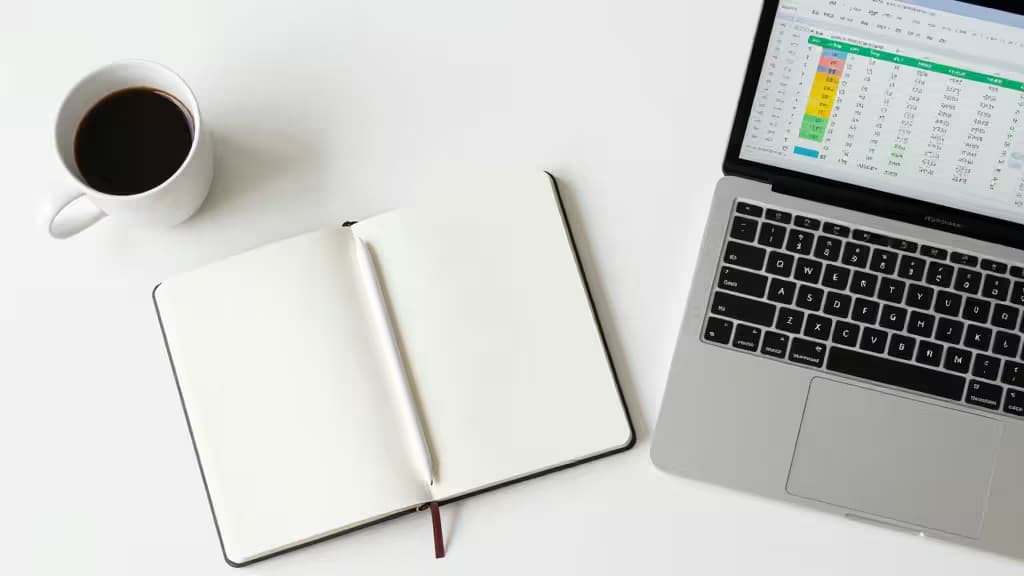 A laptop displaying expense tracking software sits beside an open notebook and coffee cup on a white desk.