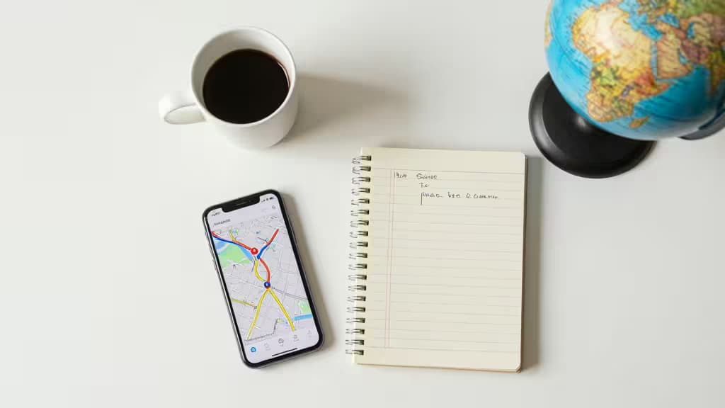 Un smartphone affichant une carte, un carnet avec des notes manuscrites, une tasse de café et un globe posés sur une table blanche.