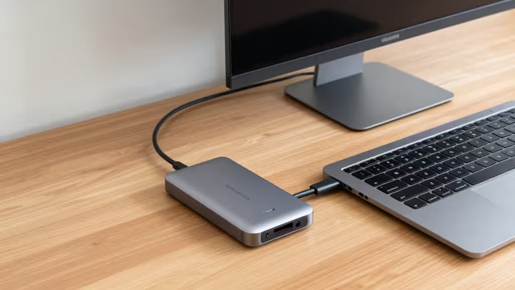 Silver USB-C hub connects MacBook Air to monitor on wooden desk.
