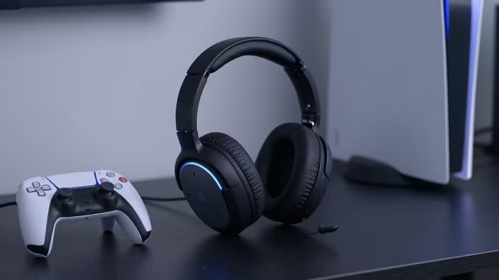 Black over-ear gaming headset with blue LED ring sits beside white PS5 controller on dark desk.