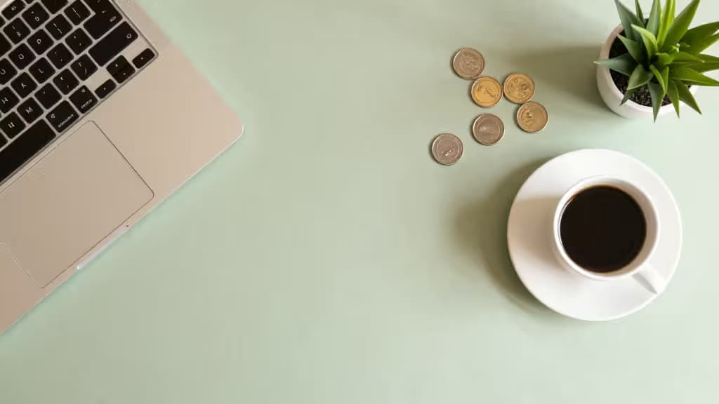 A minimalist freelancer's desk with a laptop, coffee cup, scattered coins, and a small potted plant in soft natural light.