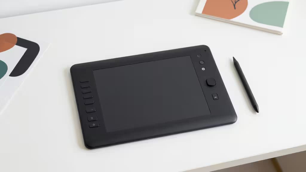 Tableta digital de dibujo negra con lápiz óptico sobre un escritorio blanco limpio, rodeada de ilustraciones abstractas.