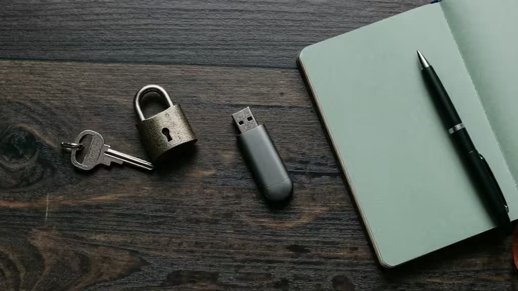 Metal key and padlock rest beside USB drive and notebook on dark wood desk.