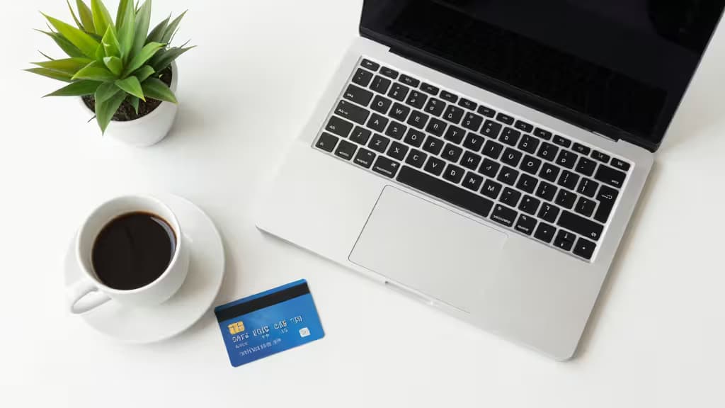 Silver laptop, blue card, white coffee cup, and green plant arranged on a white desk.