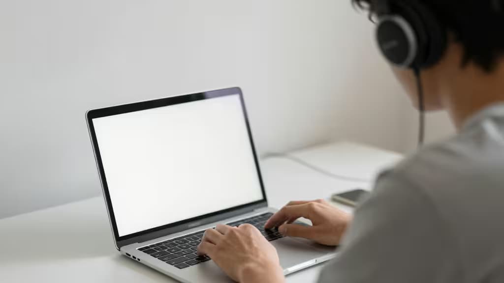 Person wearing black over-ear headphones types on silver laptop at white desk.