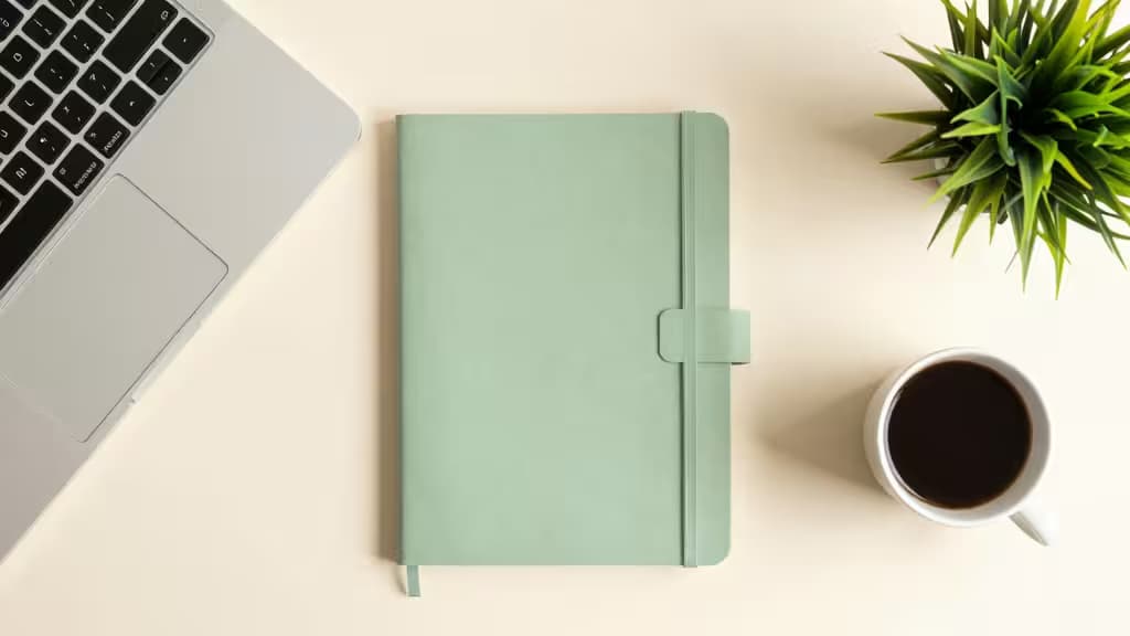 Minimalistische Schreibtischszene mit Laptop, Notizbuch, Kaffeetasse und kleiner grüner Pflanze in warmem, natürlichem Licht.