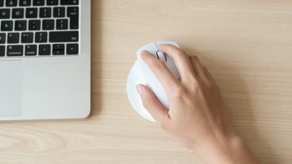 Uma mão segura um mouse vertical branco sobre uma mesa de madeira clara ao lado de um notebook prateado.
