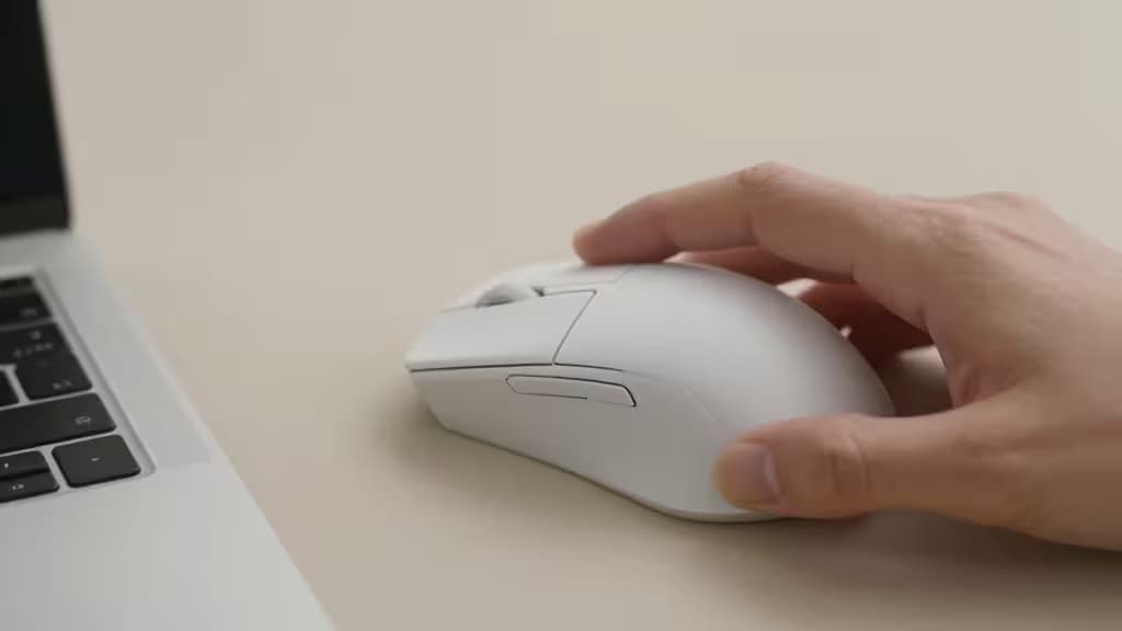 Eine Hand umfasst eine kompakte weiße kabellose Maus neben einem Laptop auf einem sauberen Schreibtisch.