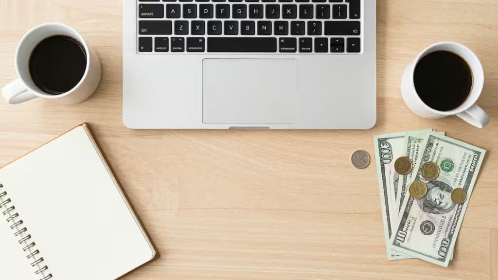 Laptop, coffee, cash, and notebook on wood desk symbolize freelance finance management.