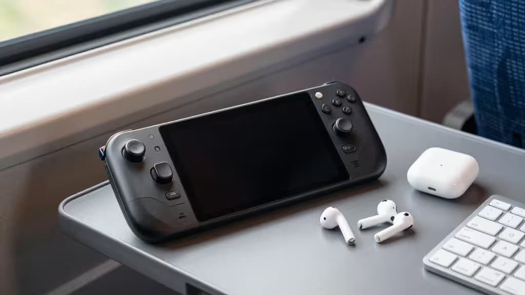 Black Steam Deck, white AirPods, and keyboard rest on a gray train tray table.