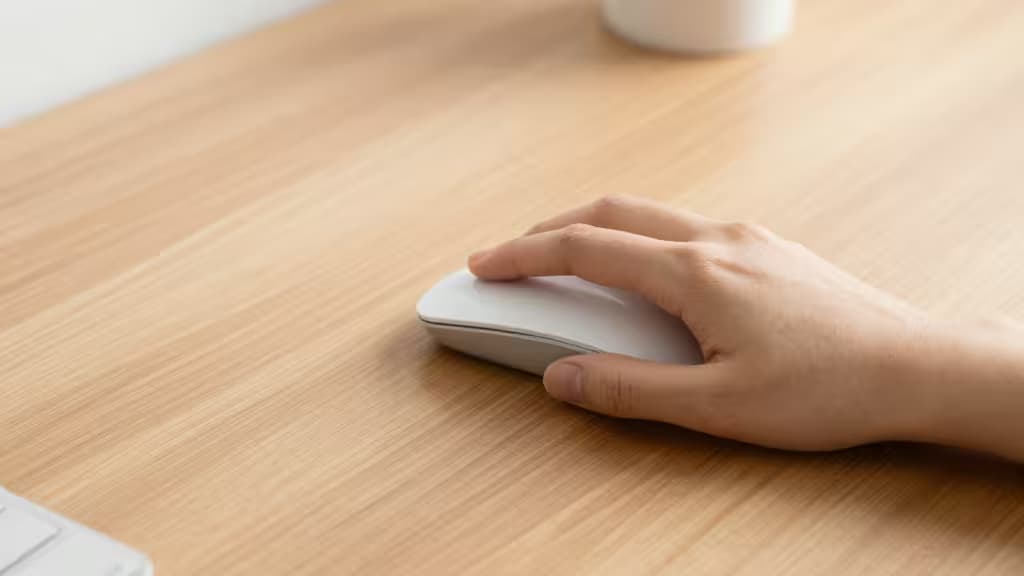 A small hand grips a compact white wireless mouse on a light wood desk.