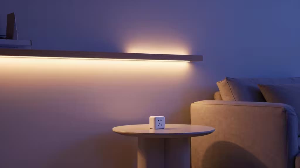 Een witte slimme stekker staat op een ronde houten tafel naast een beige bank, verlicht door warme LED-stripverlichting onder een zwevende plank.