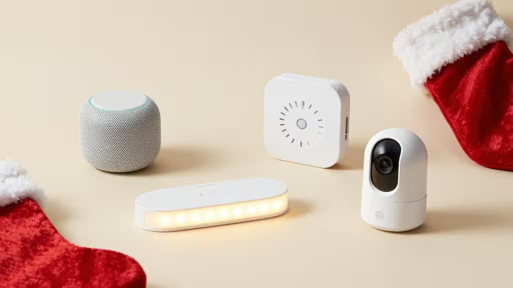 A flat-lay arrangement of small smart home gadgets including a mini speaker, water leak sensor, LED light strip, and compact security camera on a warm neutral background.