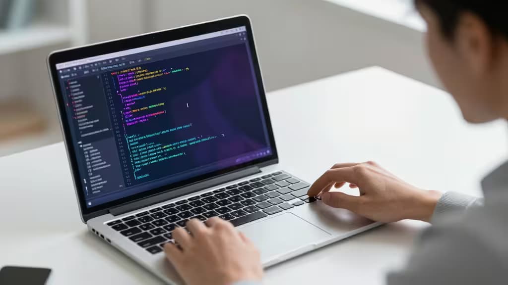 Personne tapant sur un ordinateur portable argenté affichant du code coloré, avec un arrière-plan flou évoquant un espace de travail moderne.