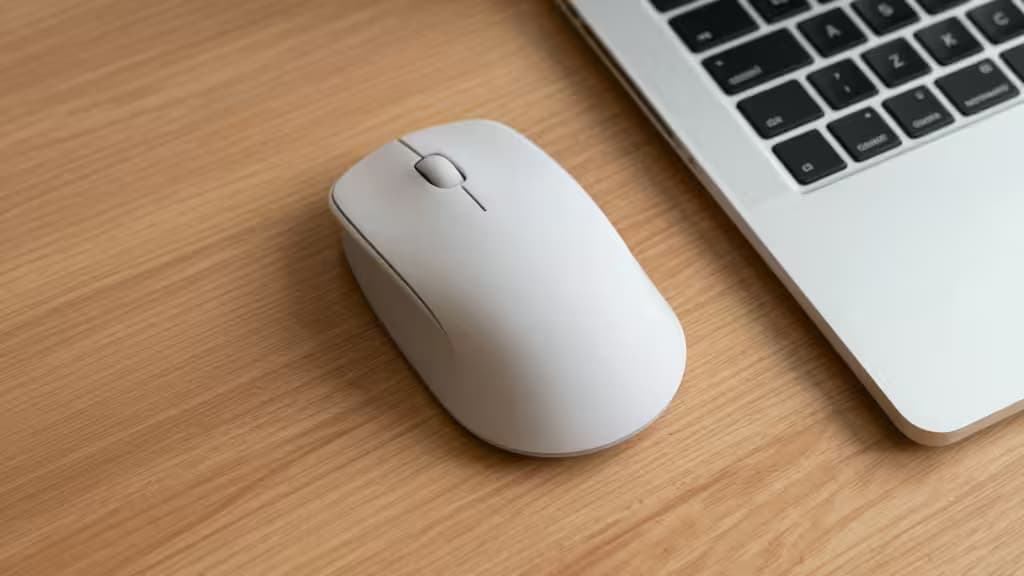 Eine weiße vertikale Maus liegt neben einem silbernen MacBook auf einem hellen Holztisch – ergonomisches Arbeiten im modernen Homeoffice