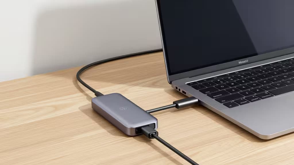 Um dock Thunderbolt 4 prateado conectado a um MacBook Pro sobre uma mesa de madeira clara, com cabos organizados e iluminação natural suave.