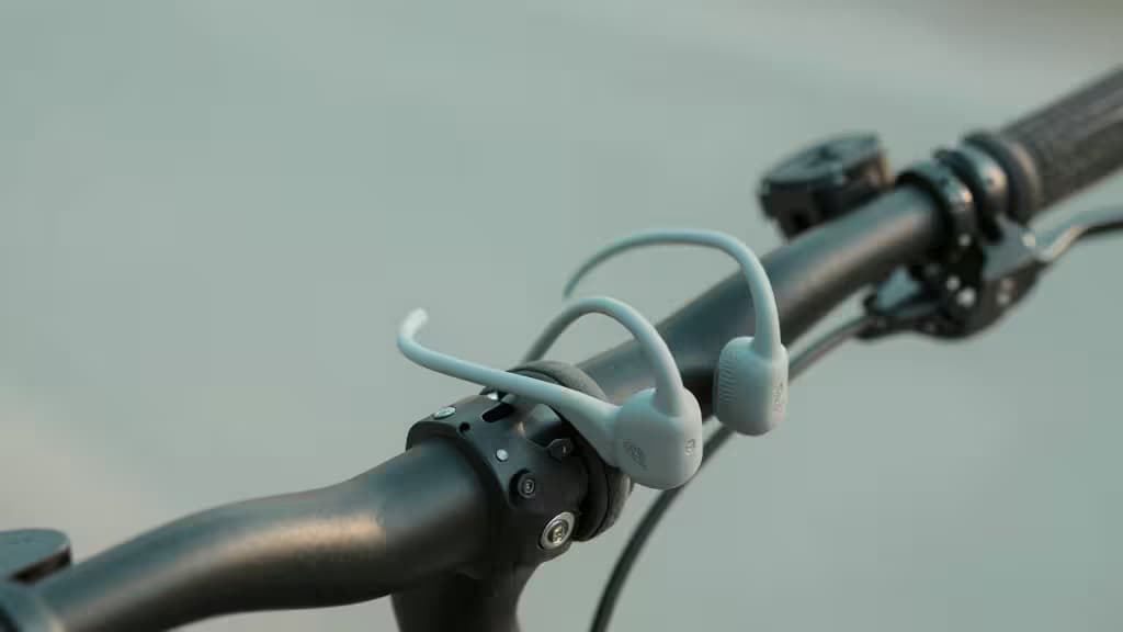 Lichtblauwe bone conduction koptelefoon rust op een matzwarte fietsstuur-stuurpen, open-ear design zichtbaar tegen een zacht uitgefocuste achtergrond.