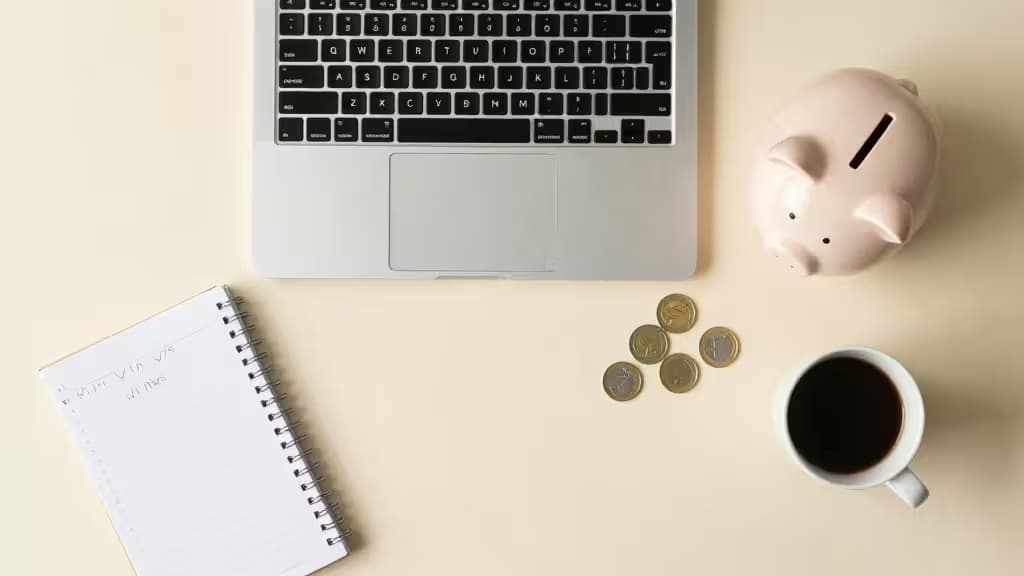Laptop, piggy bank, coins, notebook, and coffee cup arranged on beige surface.
