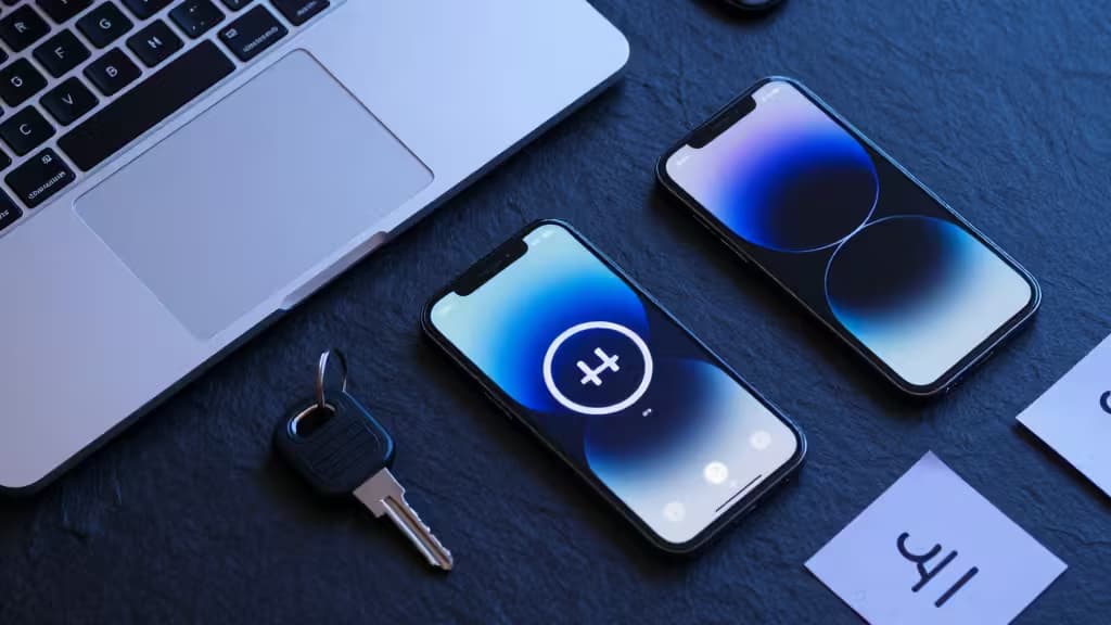 A laptop, two smartphones displaying biometric unlock screens, and a physical security key rest on a dark textured desk.