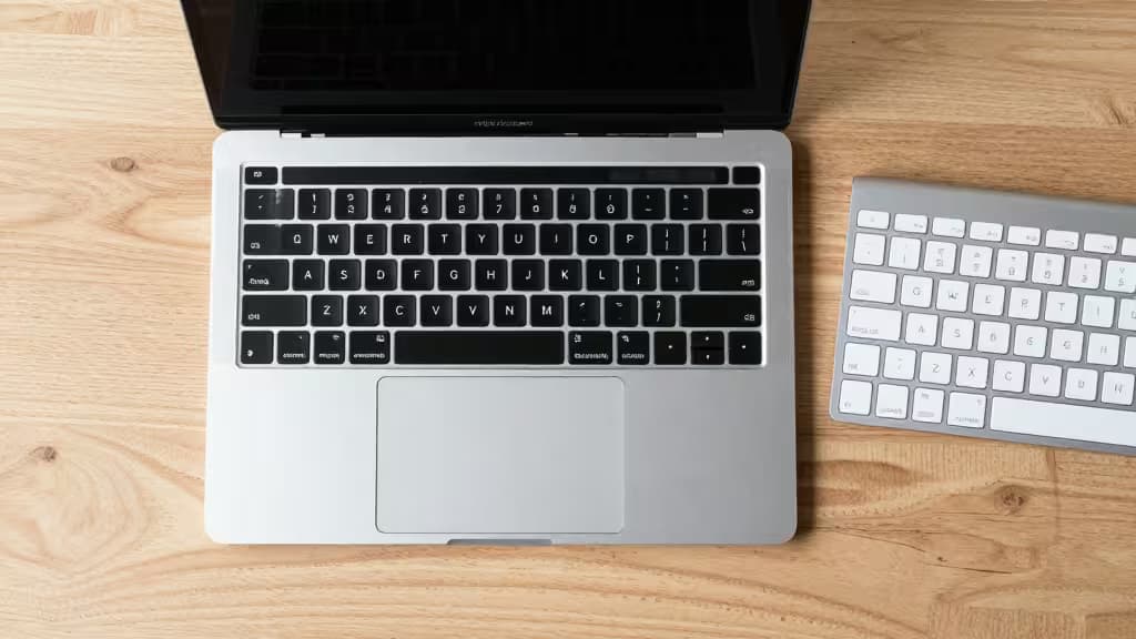 Silver MacBook with black keyboard sits beside a white wireless keyboard on light wood desk.