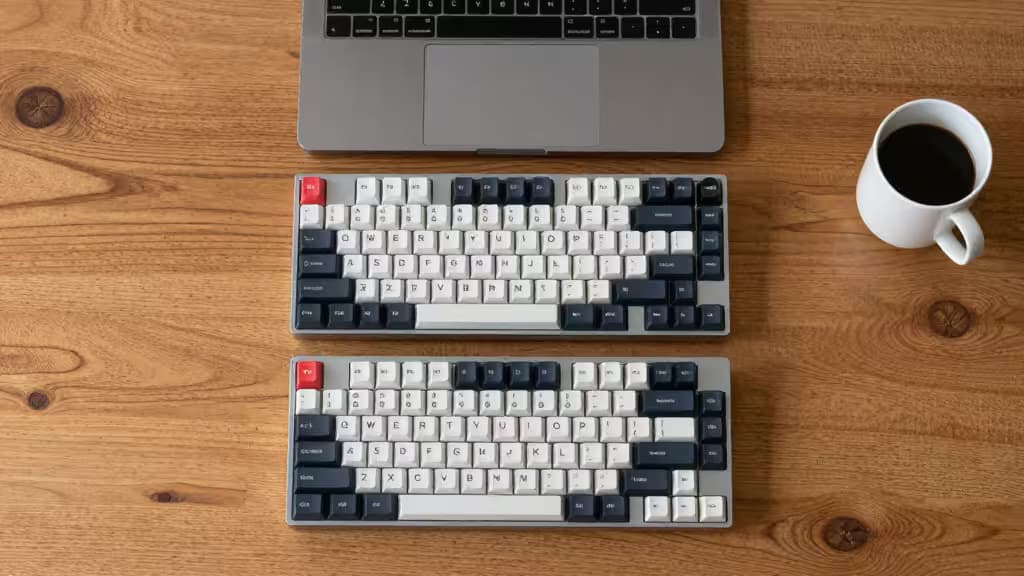 Two compact mechanical keyboards and a laptop rest on a wooden desk beside a coffee cup.