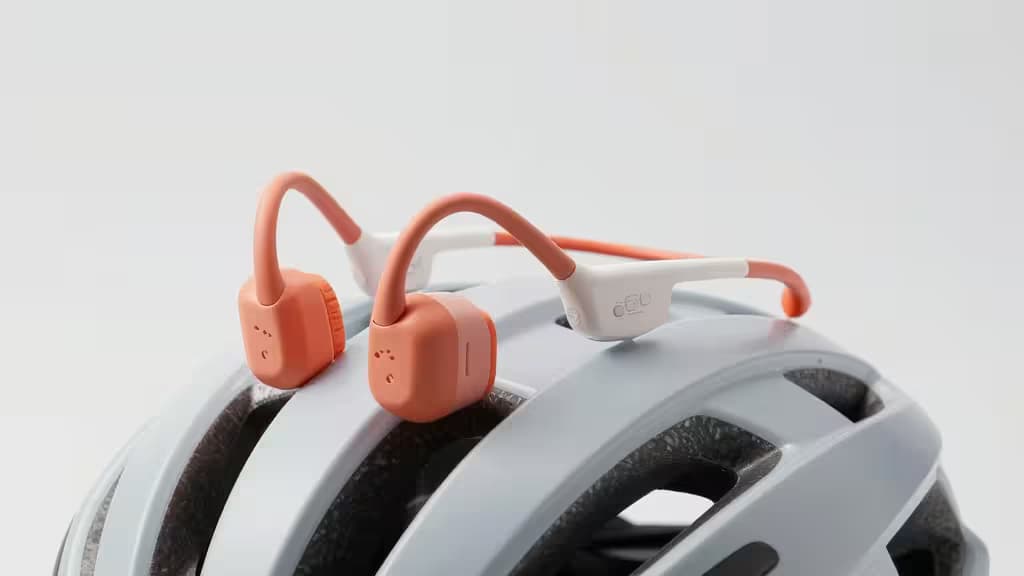 Orange and white bone conduction headphones rest atop a matte grey bicycle helmet against a clean white background.