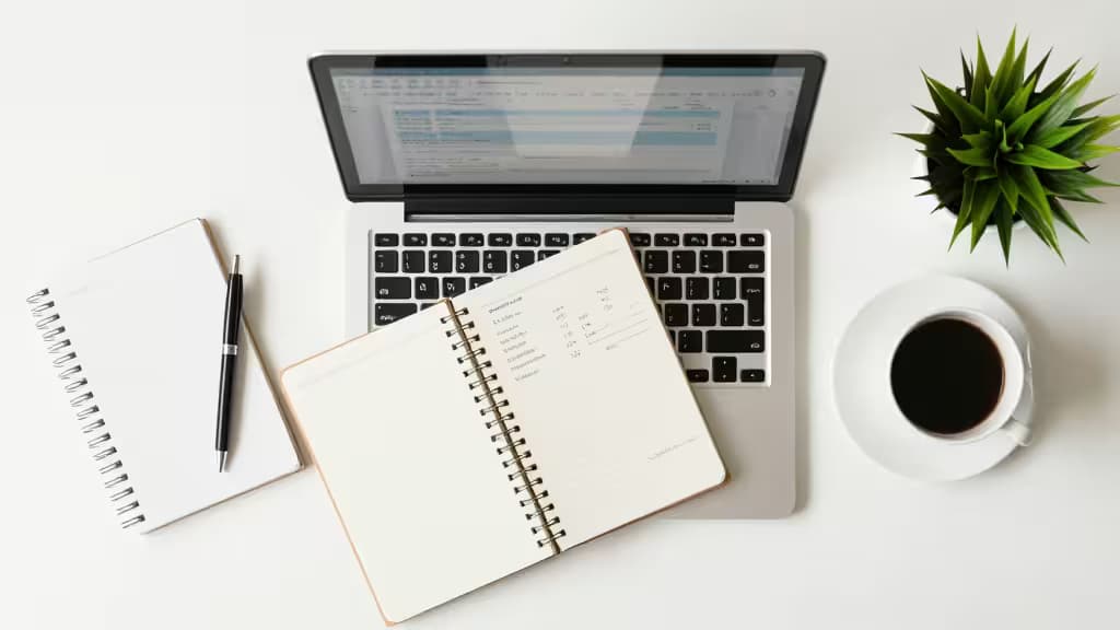 Laptop toont facturatiesoftware, omringd door notitieboekjes, pen, koffie en plant op wit bureau.