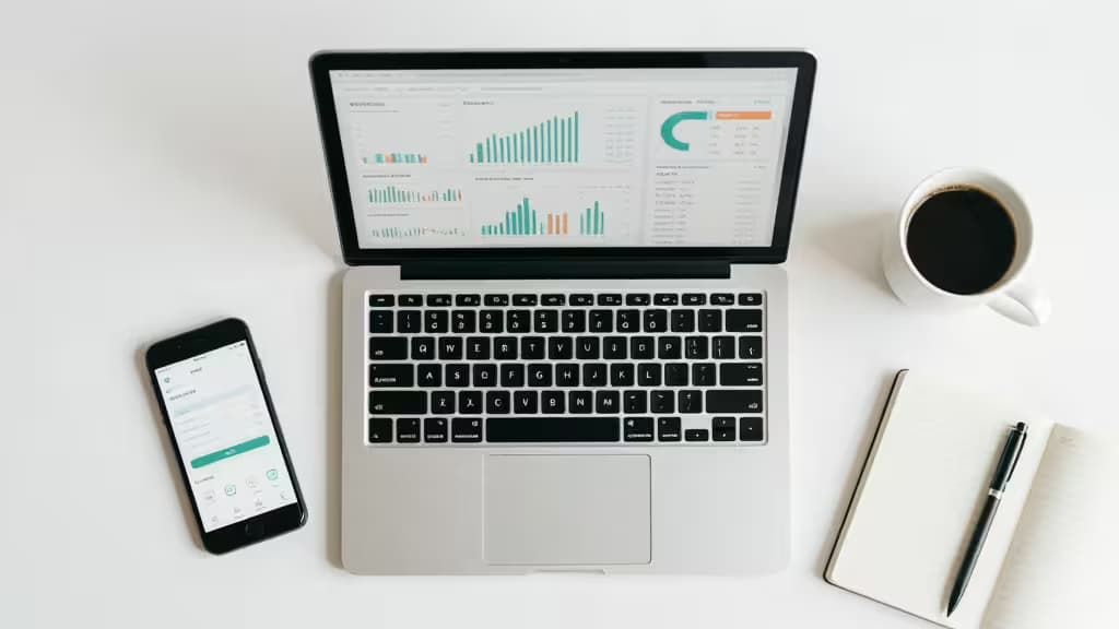 Silver laptop displays financial charts beside a phone showing banking app, notebook, pen, and coffee on white desk.