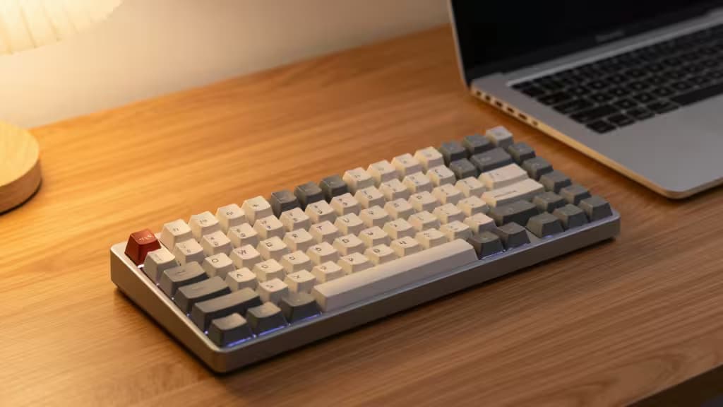 A compact mechanical keyboard with white and gray keycaps sits on a wooden desk beside a MacBook.