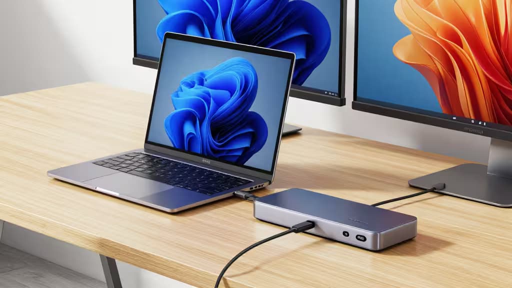 Portátil prateado ligado a um dock USB-C metálico, com dois monitores externos numa secretária de madeira clara.