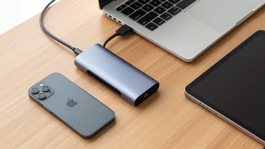 Un hub USB-C conecta un iPhone, una laptop y una tablet sobre un escritorio de madera minimalista.