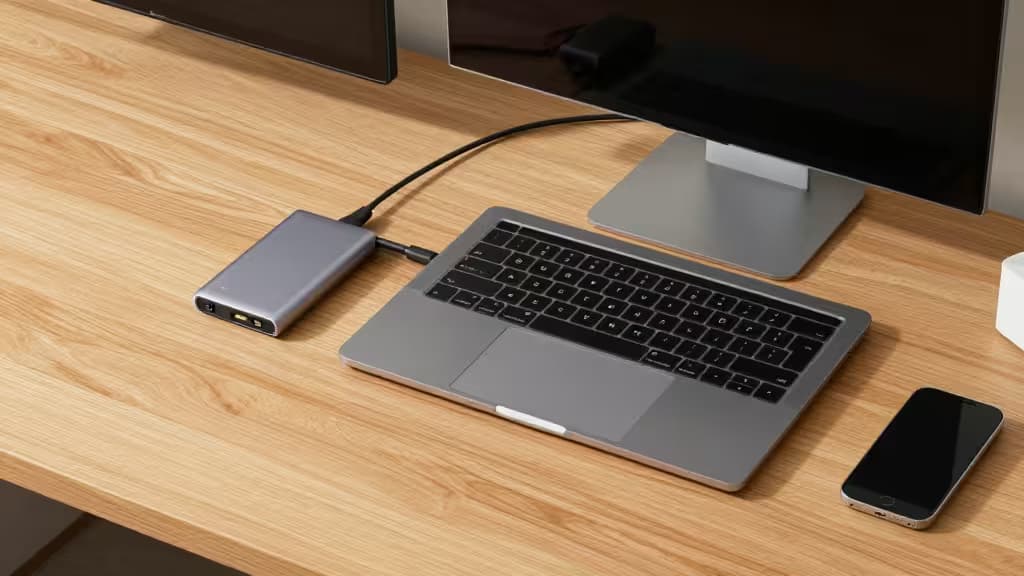 Um hub USB-C prateado e elegante conectado a um MacBook Air sobre uma mesa de madeira clara.