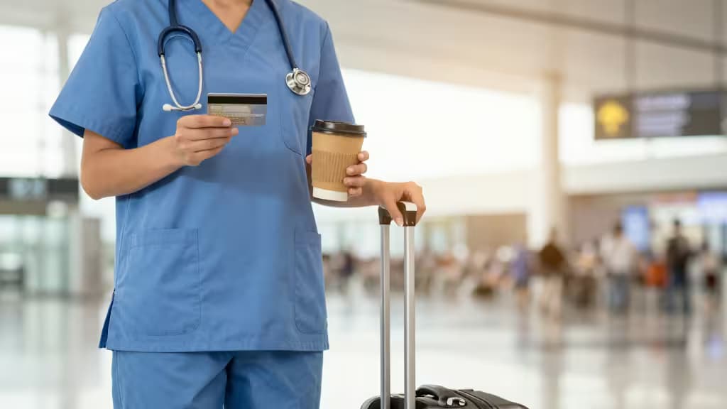 Travel nurse in blue scrubs holds credit card, coffee, and suitcase at airport terminal.