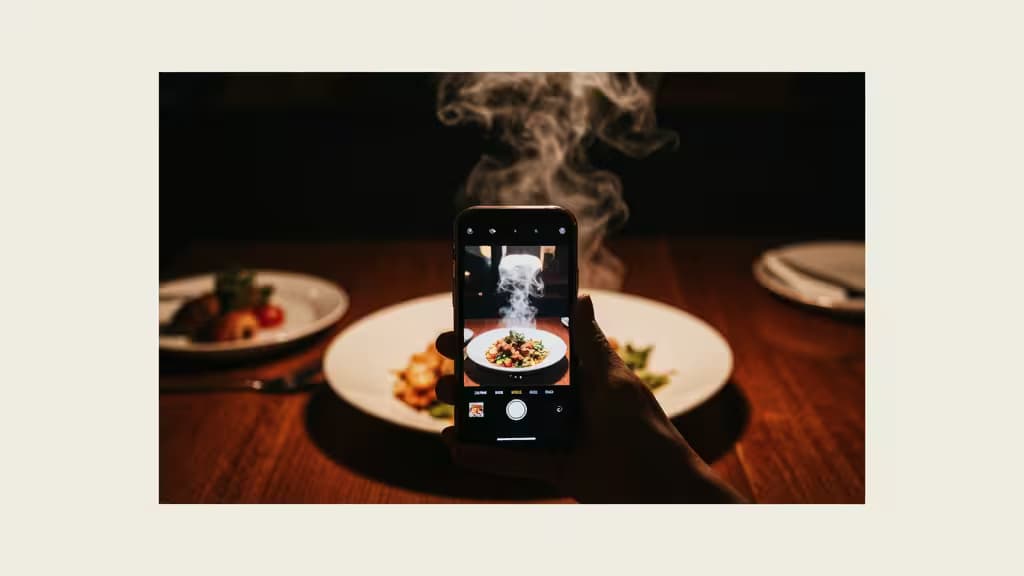 Een hand houdt een smartphone vast om een dampend gerecht op een houten tafel te fotograferen, ter illustratie van AI-foodfotografie voor restaurantmarketing.