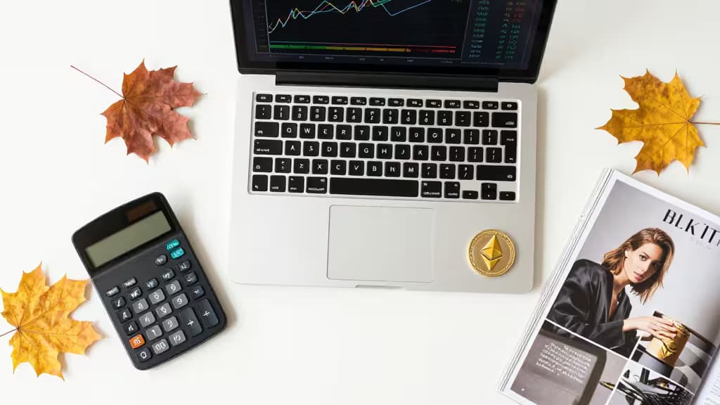 Laptop displays crypto charts, gold Ethereum coin, calculator, and autumn leaves on white desk.